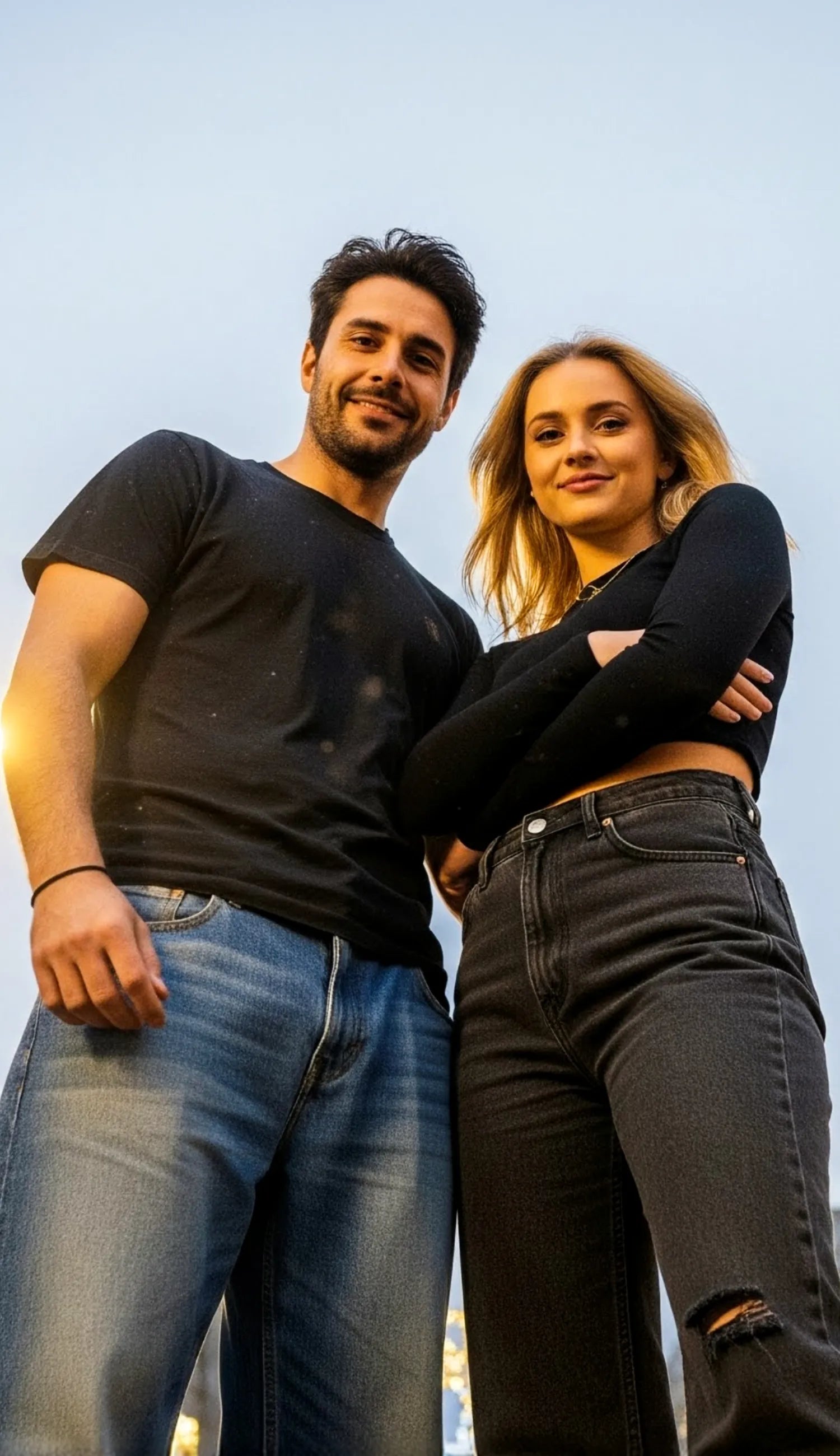 The image shows a man and woman standing confidently in an urban setting during the evening. The photo is taken from a low angle, making them appear tall and dominant against the sky. Both are wearing casual outfits—black tops and jeans. The background features modern buildings and decorative streetlights adorned with glowing string lights, giving the scene a festive yet stylish atmosphere, possibly around the holiday season or during a city event.
